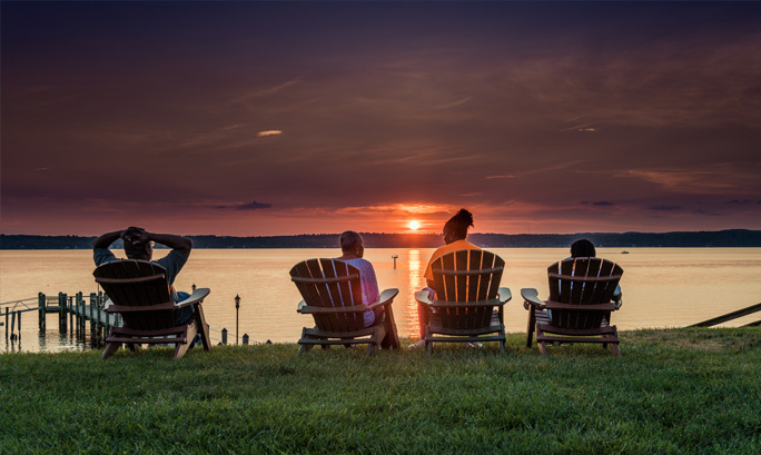 A couple sitting in Adirondack chairs watch a beautiful sunset over the lake.